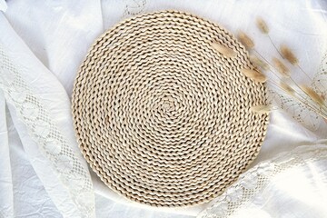 Fototapeta na wymiar Round wicker mat on linen tablecloth with lace, bohemian, rustic background mockup for product presentation, aesthetic flat lay composition.