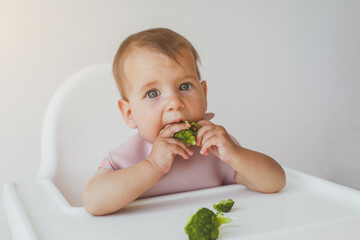 baby little girl 8 months old sits in a high chair and eats complementary foods green broccoli, close-up portrait looks at the camera