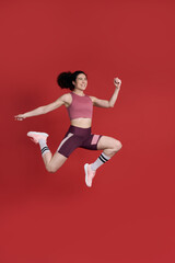 Determined young Caucasian sportswoman jumping high up, succeed training on isolated red background