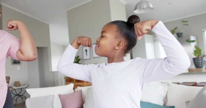 Happy unaltered african american mother and daughter flexing muscles in living room, in slow motion