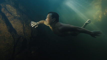 Young man free dives in the lake located in a forest and swims by underwater rocks