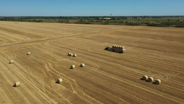 Above camera panning view, dolly move - orbit, around tractor as dragging trailer piled up with round bales of straw ride over farmland, arable field, stubble of wheat.