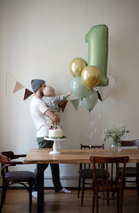 Dad with a beard and in a white T-shirt holds a little toddler boy on his shoulders against the background of festive balloons and a birthday cake