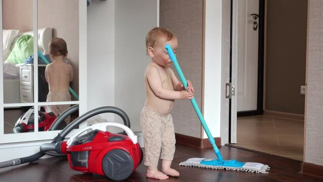 Toddler girl tries to help cleaning floor in apartment with wet mop. Daughter with bare torso in pants holding blue mop attempts doing general cleaning