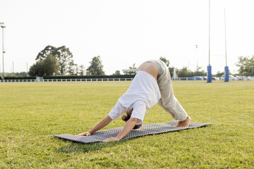 barefoot man in white t-shirt and cotton pants practicing yoga in dolphin pose on green lawn outdoors