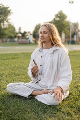 pleased man in white clothes holding aromatic palo santo stick during meditation in sukhasana pose on green grass