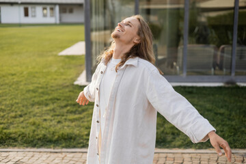 carefree yoga man in white linen shirt standing with outstretched hands and looking up near blurred house