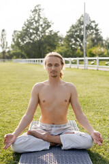Fototapeta na wymiar shirtless man in linen pants meditating in lotus pose while sitting on yoga mat with closed eyes outdoors