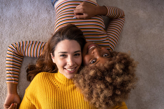 Top view of lovely multiracial lesbian couple lying on a carpet looking at camera. Beautiful happy homosexual women smiling and relaxing together on the floor.