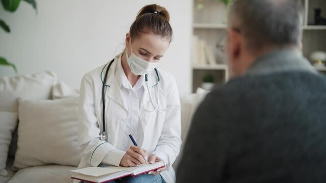 Empathic woman doctor holding notepad and laptop consulting senior man in modern private clinic. Medical practitioner ask questions, filling disease history form, provide help prescribe treatment.