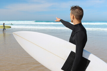 Surfer men in swimsuit holding white surfboard and pointing on the ocean waves on background. Water sport surfing concept