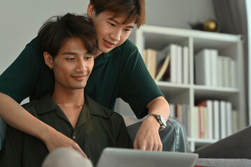 Fototapeta na wymiar Happy same sex male couple embracing and using laptop in living room. LGBT, love and lifestyle relationship concept