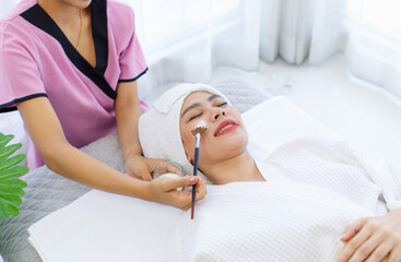 Millennial Asian professional unrecognizable female masseuse therapist in uniform standing using brush brushing essential oil cream on customer face while laying down closed eyes relax on massage bed