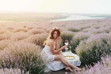Young curly woman in white dress drinking wine in lavender field. Picnic on the field.	