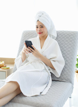 Millennial Asian female customer in white clean bathrobe and towel sitting on armchair playing with smartphone waiting for masseuse do honey wax aromatherapy massaging