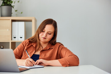 An ambitious curvy businesswoman writing notes and working on a laptop.