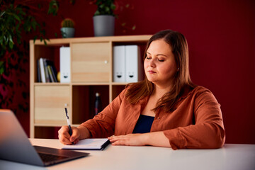 A pretty plus-size businesswoman writing notes while working over a laptop.