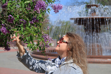 Attractive woman in sunglasses enjoys the smell of lilacs in a city park.