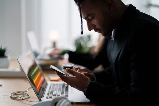 Young black man using smartphone while checking messages during work on laptop in modern workspace