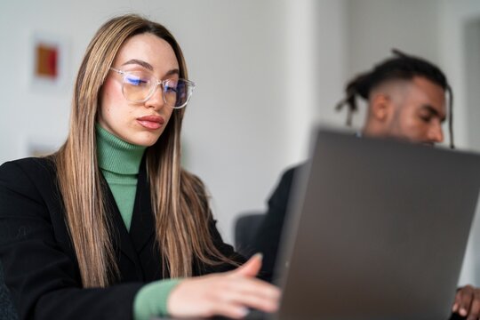 Concentrated young female entrepreneur in formal wear and eyeglasses typing information on laptop keyboard while working on business strategy together with African American coworker in modern office