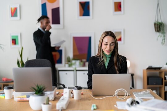 Positive businesswoman in green sweater and black blazer sitting at table with takeaway coffee and using laptop while looking at camera during workday in office