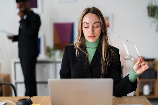 Positive businesswoman in green sweater and black blazer sitting at table with takeaway coffee and using laptop while looking at camera during workday in office