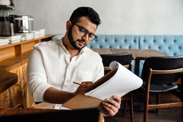 Handsome indian male business owner in eyeglasses working with paper documents in cafe