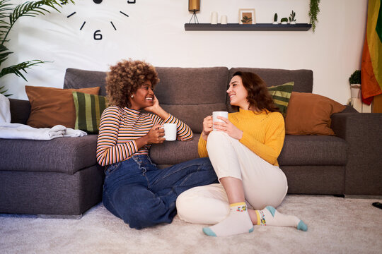 Happy young lesbian couple relaxing together in their living room at home. Smiling female lovers spending quality time together with coffees and talks. Full length.
