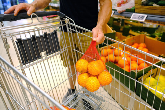 male shopper holding in hand orange fruits in plastic bag, holds ripe orange in hand, puts groceries in iron grocery trolley, healthy eating, vegan diet, vitamin c, industrial citrus fruit production