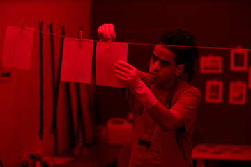 Young photographer hanging printed photos on rope in darkroom with red lighting
