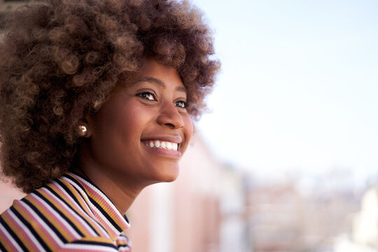 Happy young black woman with afro hair smiling. Cheerful African American girl watching outdoors in open air. Portrait of female with positive expression looking out of window. Copy space.