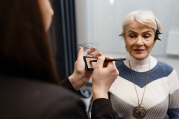 An elderly woman visiting a professional makeup artist. An experienced makeup artist does makeup for a client