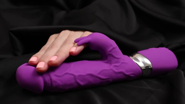 A woman's hand takes a purple dildo on a black silk sheet. 