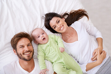 Fototapeta premium Top view, portrait and happy parents with baby on bed for love, care and quality time together at home. Smile of mother, father and family with cute newborn kid relaxing in bedroom, fun and happiness