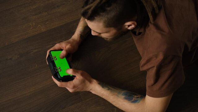 Caucasian man using smartphone in horizontal mode with green mock-up screen is lying on a wooden floor and playing online games. Guy uses a joystick for phone. Focus on Chroma Display. Top view.