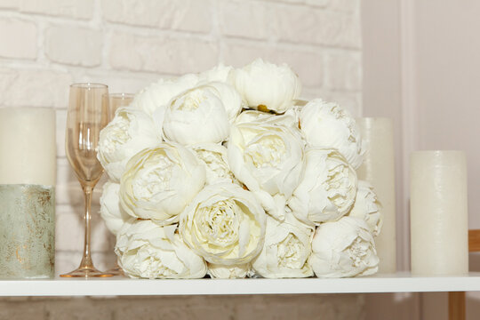 A wedding white bouquet of peonies, glasses and white candles lie on shelf. Beautiful wedding set for bride