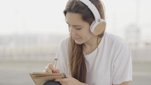Young woman listening music while writing on notebook outside
