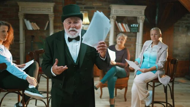 Senior man actor with plenty of facial expressions rehearsing his lines from a script and getting applause from fellow actors on stage
