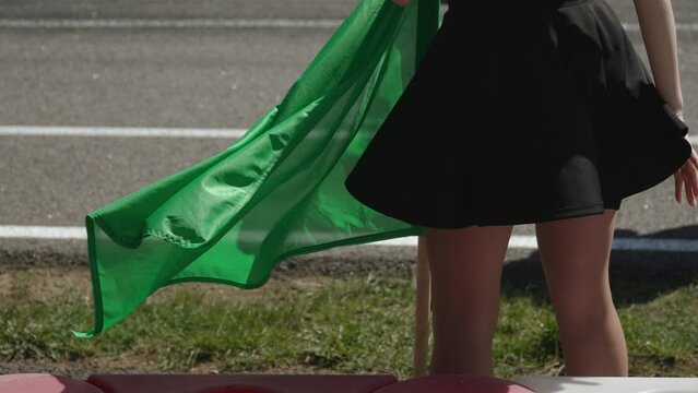 Young woman with green flag flying near starting grid of race track has her skirt lifted by wind and reveals her buttocks in pantyhose. Operation of model for signaling start of race.