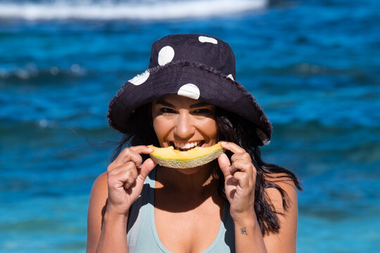 Close up of Young woman wearing full brim hat and eating rockmelon on the beach on clear sunny day