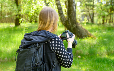 Kobieta z aparatem fotograficznym na szyi ogląda zrobione zdjęcia na ekranie lustrzanki stojac w lesie 