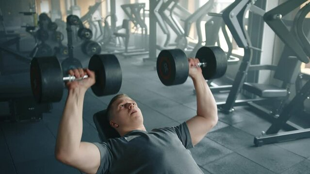 Medium shot of young sportsman performing dumbbell bench press in equipped gym. Man lies on bench, stretches arms vertically and lowers dumbbells down. High quality 4k footage
