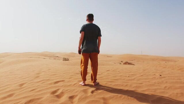 Barefoot young man stands in desert and looks around. Back view. Concept of relaxation and contemplation. Slow motion