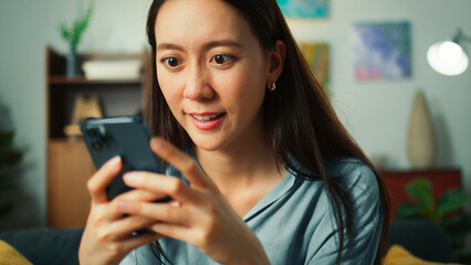 Fototapeta na wymiar Closeup of Happy beautiful Asian woman sit on sofa hand holding mobile phone chatting and playing social media. Young female looking at smartphone cellphone browsing internet on sofa home living room