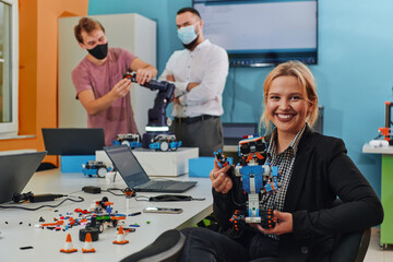 A woman sitting in a laboratory and solving problems and analyzing the robot's verification. In the background, colleagues are talking at an online meeting