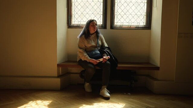 Portrait of overweight young woman sitting on bench under windows of Paris Musee de Cluny in warm sunlight, examining exhibition. Caucasian female visits exhibition of Musee National du Moyen Age.