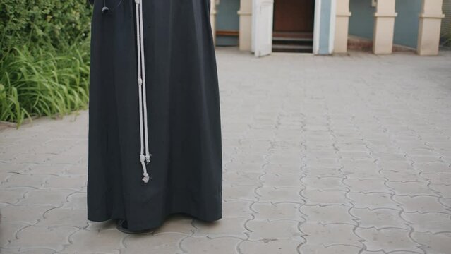 Close-up of the lower body of church minister. Franciscan priest in black long cassock tied with white rope in the parish courtyard