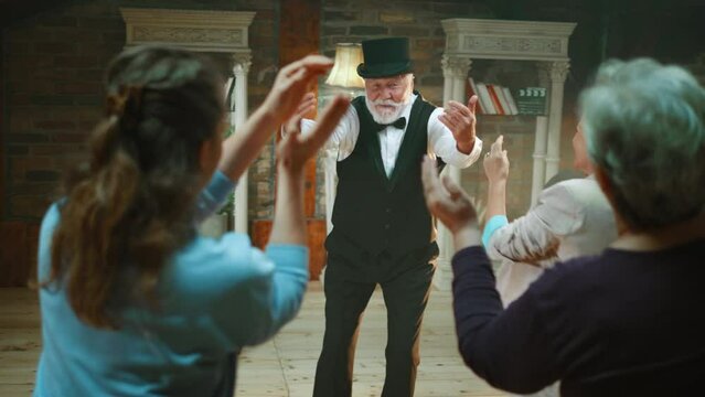 Cheerful elderly male comedian, presenter or actor invites the audience to join him on stage and dance to the music during the performance on the small stage
