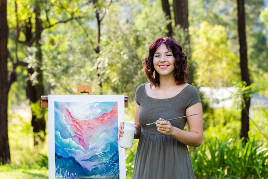 Happy young australian artist holding white paint pot and brush beside artwork