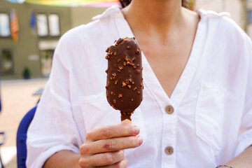 an unrecognizable woman holds a almond-shaped chocolate lolly at a town festival. ice cream in summer - obrazy, fototapety, plakaty
